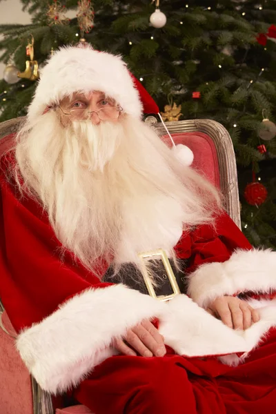 Санта-Клаус сидит в кресле перед елкой — стоковое фото