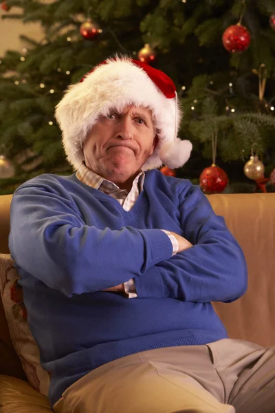 Seniorchef als Weihnachtsvater verkleidet sieht mürrisch vor dem Weihnachtsbaum aus — Stockfoto