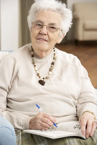 Senior vrouw ontspannen in stoel thuis voltooiing kruiswoordraadsel — Stockfoto