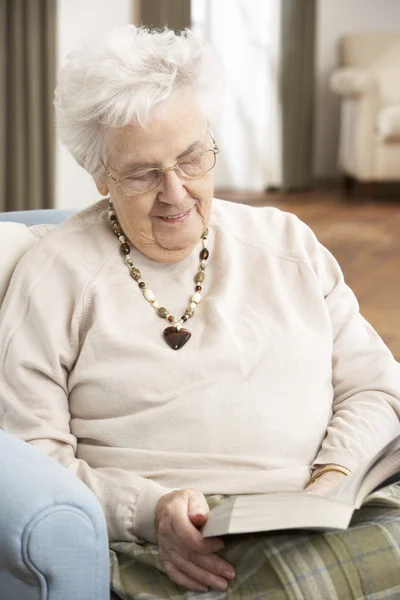 Senior mulher relaxante na cadeira em casa leitura livro — Fotografia de Stock