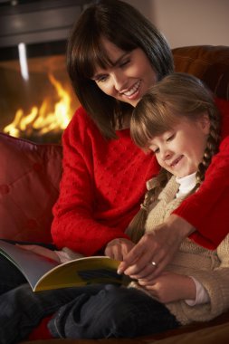 Mother And Daughter Sitting On Sofa And Reading Book By Cosy Log clipart