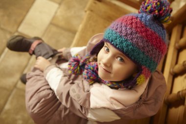 Young Girl Sitting On Wooden Seat Putting On Warm Outdoor Clothe clipart