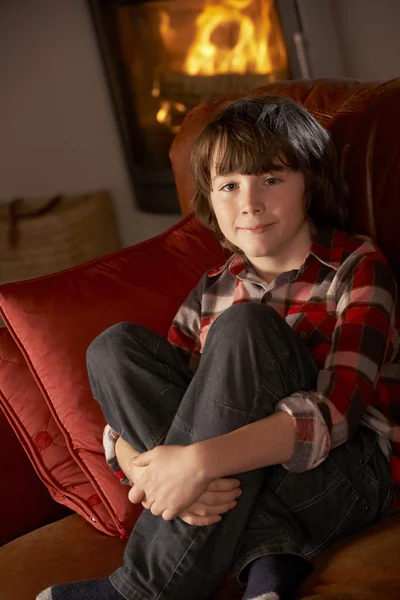 Young Boy Sitting On Sofa By Cosy Log Fire — Zdjęcie stockowe