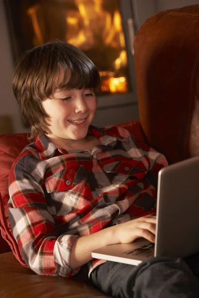 Chico joven que relaja con el ordenador portátil por el fuego de registro acogedor —  Fotos de Stock