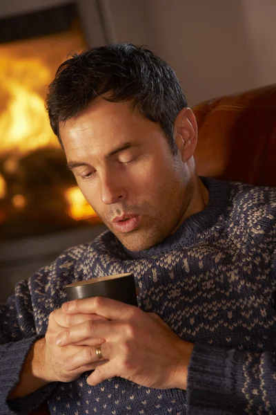 Średniej wieku człowiek relaksujący z gorącego napoju przez przytulnym kominkiem — Zdjęcie stockowe