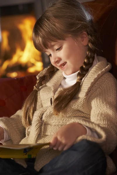 Chica joven sentada en el sofá y libro de lectura por fuego de registro acogedor — Foto de Stock