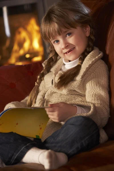Junges Mädchen sitzt auf Sofa und liest Buch am gemütlichen Kaminfeuer — Stockfoto