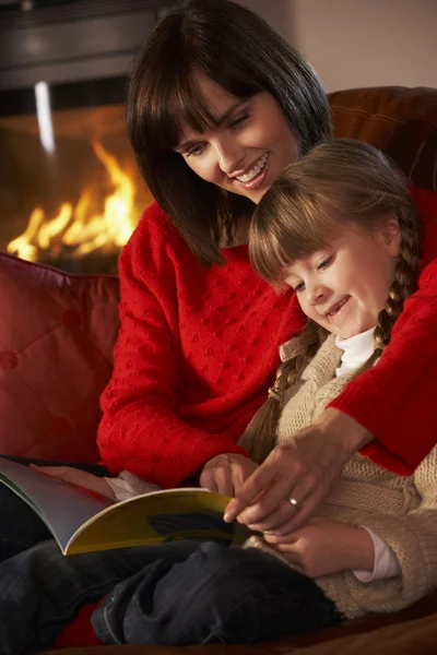 Madre e hija sentadas en el sofá y leyendo el libro por acogedor registro — Foto de Stock