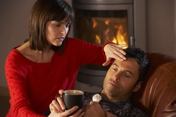 Wife Nursing Sick Husband With Cold Resting On Sofa By Cosy Log — Stock Photo, Image