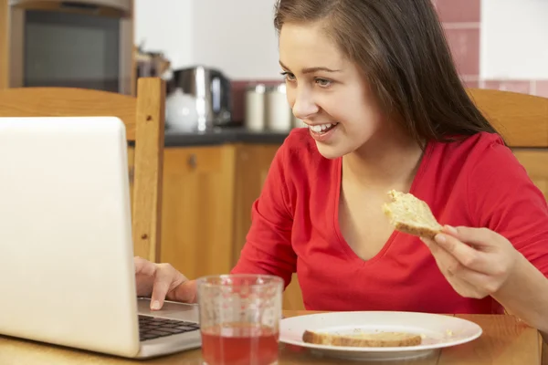 Adolescente chica usando el ordenador portátil mientras come desayuno — Foto de Stock