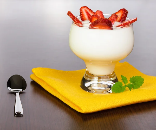 stock image Yogurt with strawberry slices in a kremanka on a yellow napkin with a spoon, located on a brown table with a wood structure