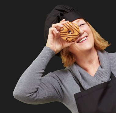 Middle aged cook woman looking through a donut over black backgr clipart