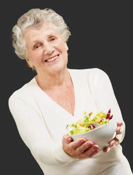 Portrait of senior woman showing a fresh salad over black backgr — Stock Photo, Image