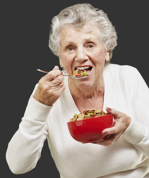Senior woman eating cereals out of a red bowl against a black ba — Stock Photo, Image