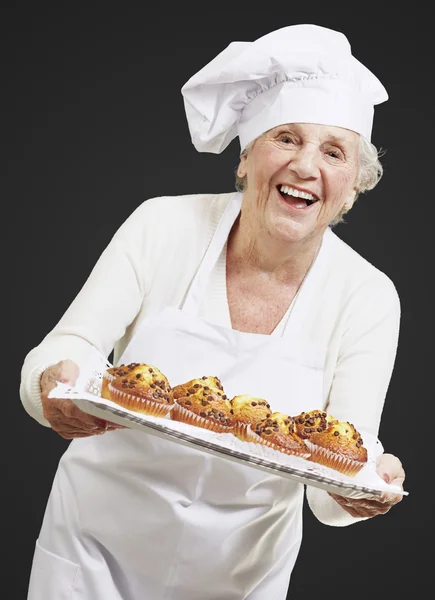 Mulher sénior cozinheiro segurando uma bandeja com muffins contra um ba preto — Fotografia de Stock