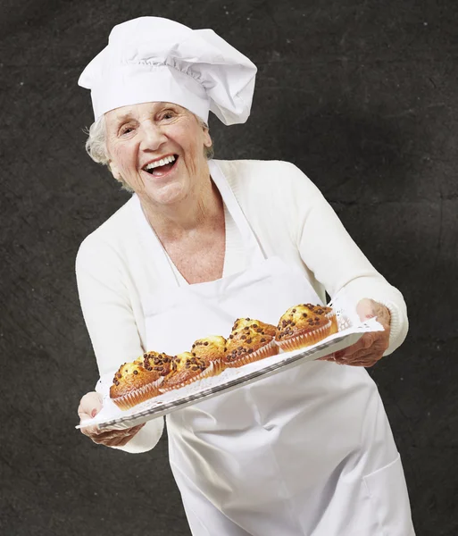 Bir tepsi kekleri ile karşı bir grunge b holding üst düzey kadın aşçı — Stok fotoğraf
