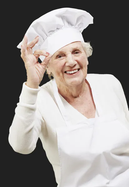 Äldre kvinna kock gör en utmärkt symbol mot en svart baksida — Stockfoto