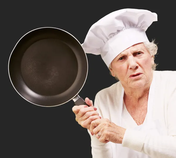 Cook senior woman angry trying to hit with pan over black backgr — Stock Photo, Image