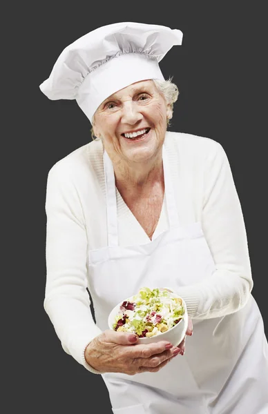 La cuoca anziana cucina tenendo una ciotola con insalata contro un dorso nero — Foto Stock