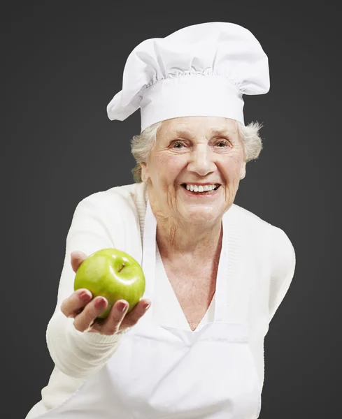 Mulher sénior cozinhar oferecendo uma maçã verde contra um backgro preto — Fotografia de Stock