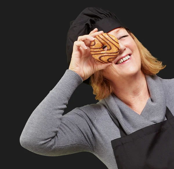 Mulher cozinheira de meia idade olhando através de um donut sobre backgr preto — Fotografia de Stock