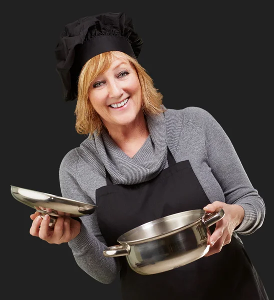 Mulher cozinheira de meia idade segurando uma panela de souce sobre fundo preto — Fotografia de Stock