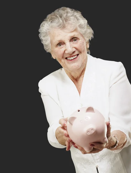 Porträt einer Seniorin, die ein Sparschwein über schwarzem Hintergrund zeigt — Stockfoto