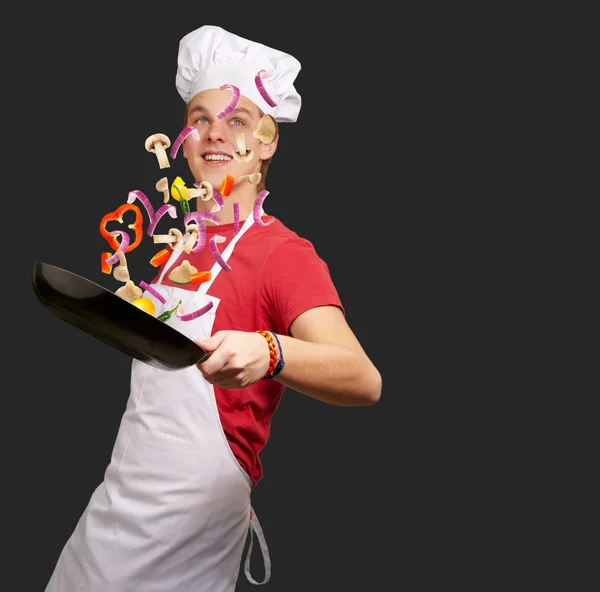 Портрет молодого кухаря, який готує овочі на сковороді над чорним — стокове фото