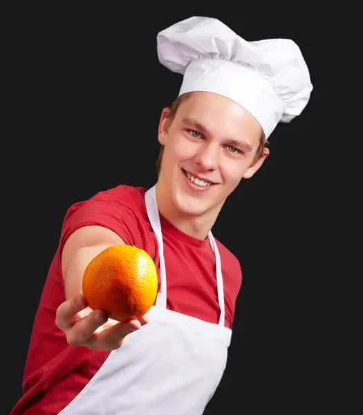 Portrait of young cook man offering orange over black background — Stock Photo, Image