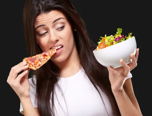 Retrato de una mujer joven comiendo pizza y buscando ensalada sobre blac —  Fotos de Stock