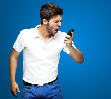 Portrait of angry young man shouting using mobile over blue background clipart