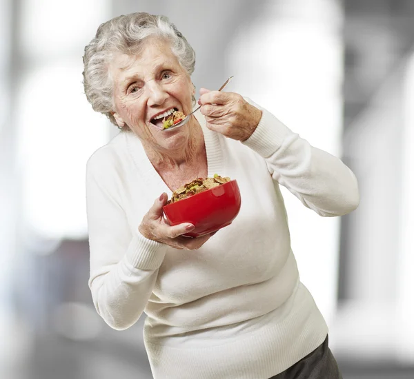 Seniorin isst Müsli aus einer roten Schüssel, drinnen — Stockfoto