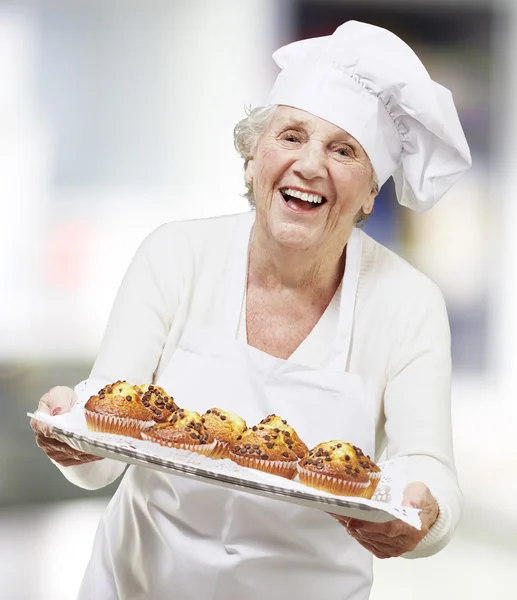 Μάγειρας ανώτερος γυναίκα, κρατώντας ένα δίσκο με muffins, εσωτερική — Φωτογραφία Αρχείου