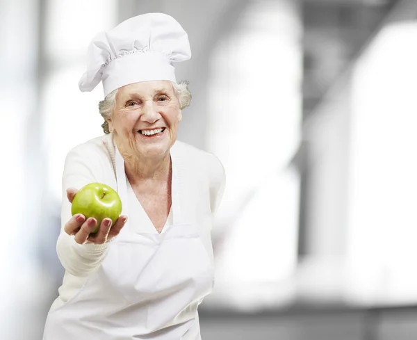 Senior woman cook offering a green apple, indoor — Stock Photo, Image