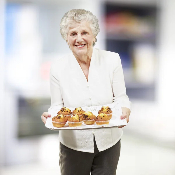 Пожилая женщина улыбается и держит поднос с кексами, в помещении — стоковое фото