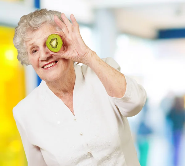 Portrait of senior woman holding kiwi in front of her eye indoor — Stock Photo, Image