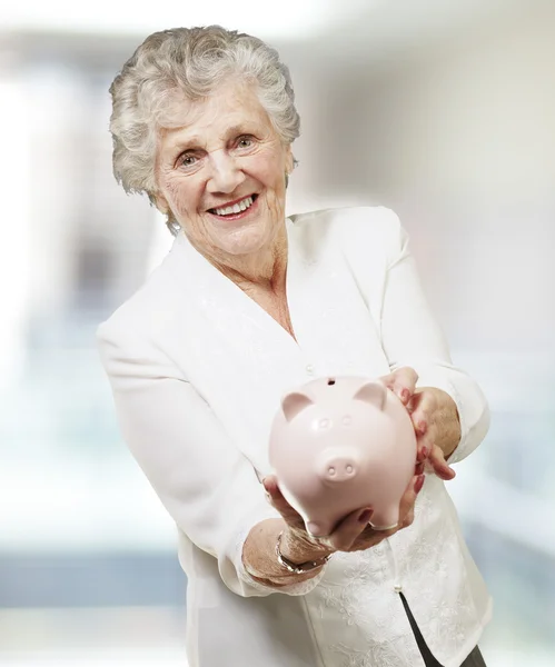 Retrato de mulher sênior mostrando um porquinho banco, indoor — Fotografia de Stock