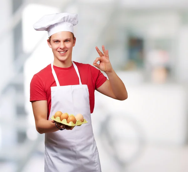 Portrait d'un jeune cuisinier tenant une boîte à œufs et faisant un bon gestur — Photo