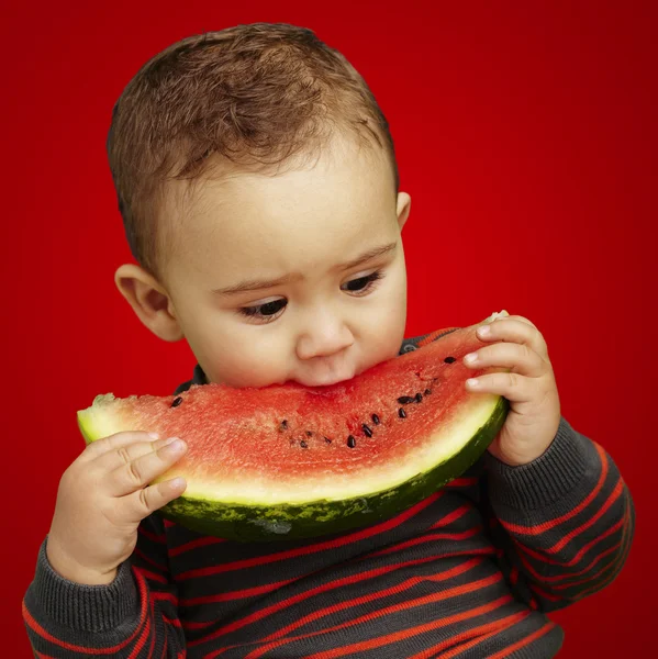 Portret przystojny dziecko gryzie arbuza na czerwonym tle — Zdjęcie stockowe