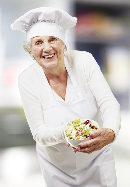 Mulher sênior cozinheiro segurando uma tigela com salada, interior — Fotografia de Stock