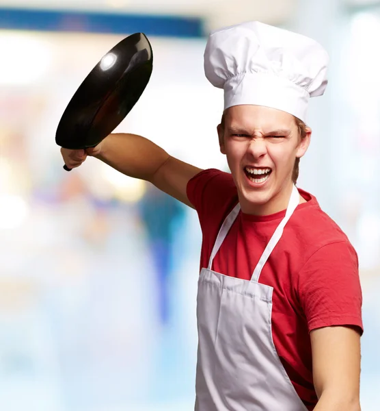 Portrait of angry young cook man hitting with pan indoor — Stock Photo, Image