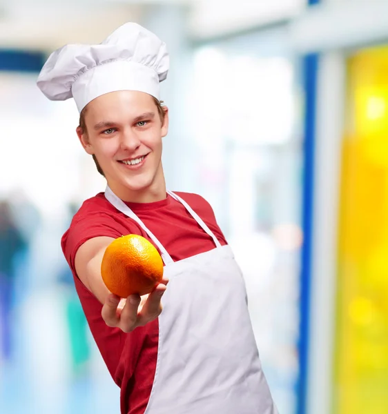Portrait of young cook man offering orange indoor — Stock Photo, Image