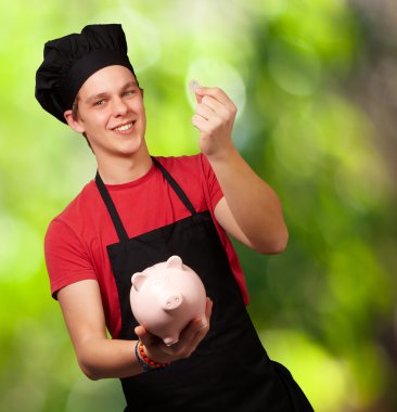 Portrait of young cook man holding euro coin and piggy bank agai