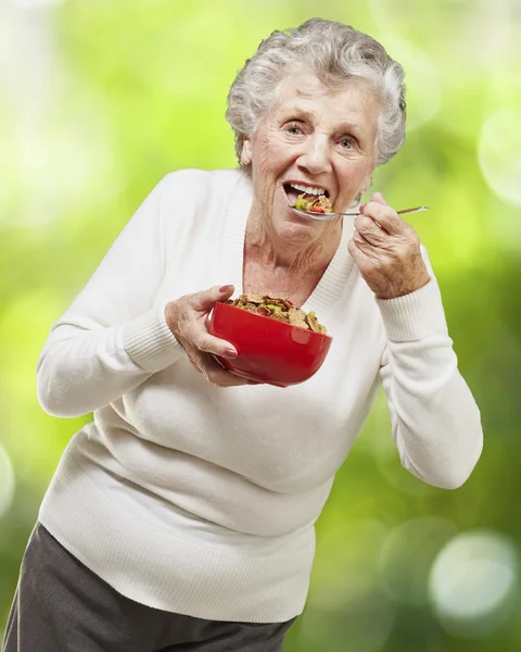 Senior woman eating cereals out of a red bowl against a nature b — Stock Photo, Image