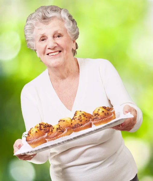 Portrait of senior woman showing a chocolate muffin tray against — Stock Photo, Image