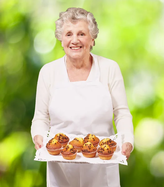 Retrato de cocinera mujer mayor sosteniendo una bandeja de magdalenas de chocolate a —  Fotos de Stock