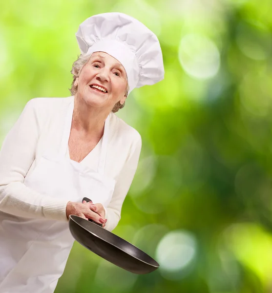 Portrait of a friendly cook senior woman holding pan against a n — Stock Photo, Image