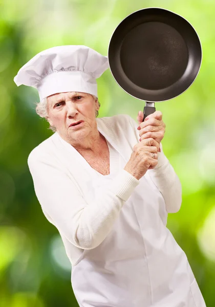 Cook senior woman angry trying to hit with pan against a nature — Stock Photo, Image