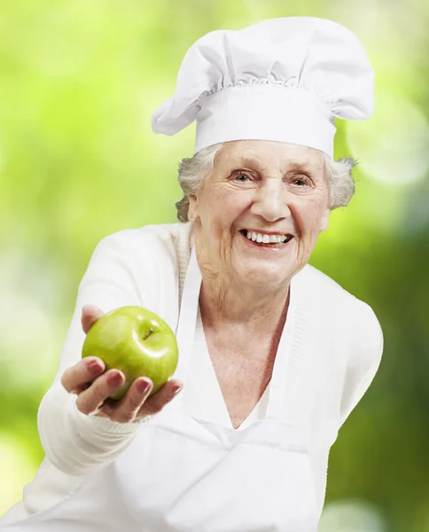 Senior woman cook offering a green apple against a nature backgr — Stock Photo, Image