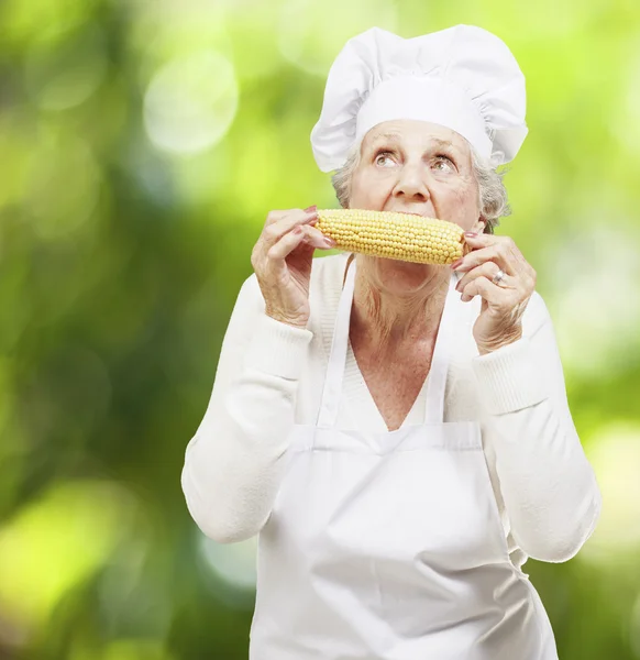 Donna anziana cuoco mangiare una pannocchia di mais su uno sfondo naturale — Foto Stock